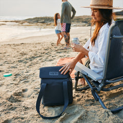 Navy/ 14L | YETI Daytrip Insulated Tote Bag Image Showing Tote Bag On The Sand Next To A Model, Being Closed After Taking A Can Of Drink Out Of The Tote Bag.