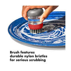 OXO Outdoor Soap Dispensing Brush With Storage Case | Image Showing The Brush Scrubbing A Camp Plate.