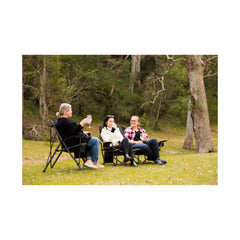 Black | Oztrail Fireside Double Chair Image Showing Two Models Sitting In Chair At Camp.