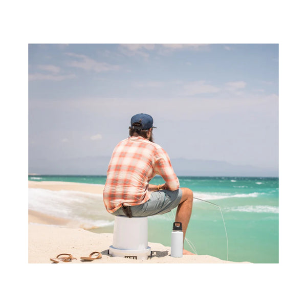 White | YETI Loadout Bucket Image Showing A Model Using The Bucket As A Seat While Beach Fishing.